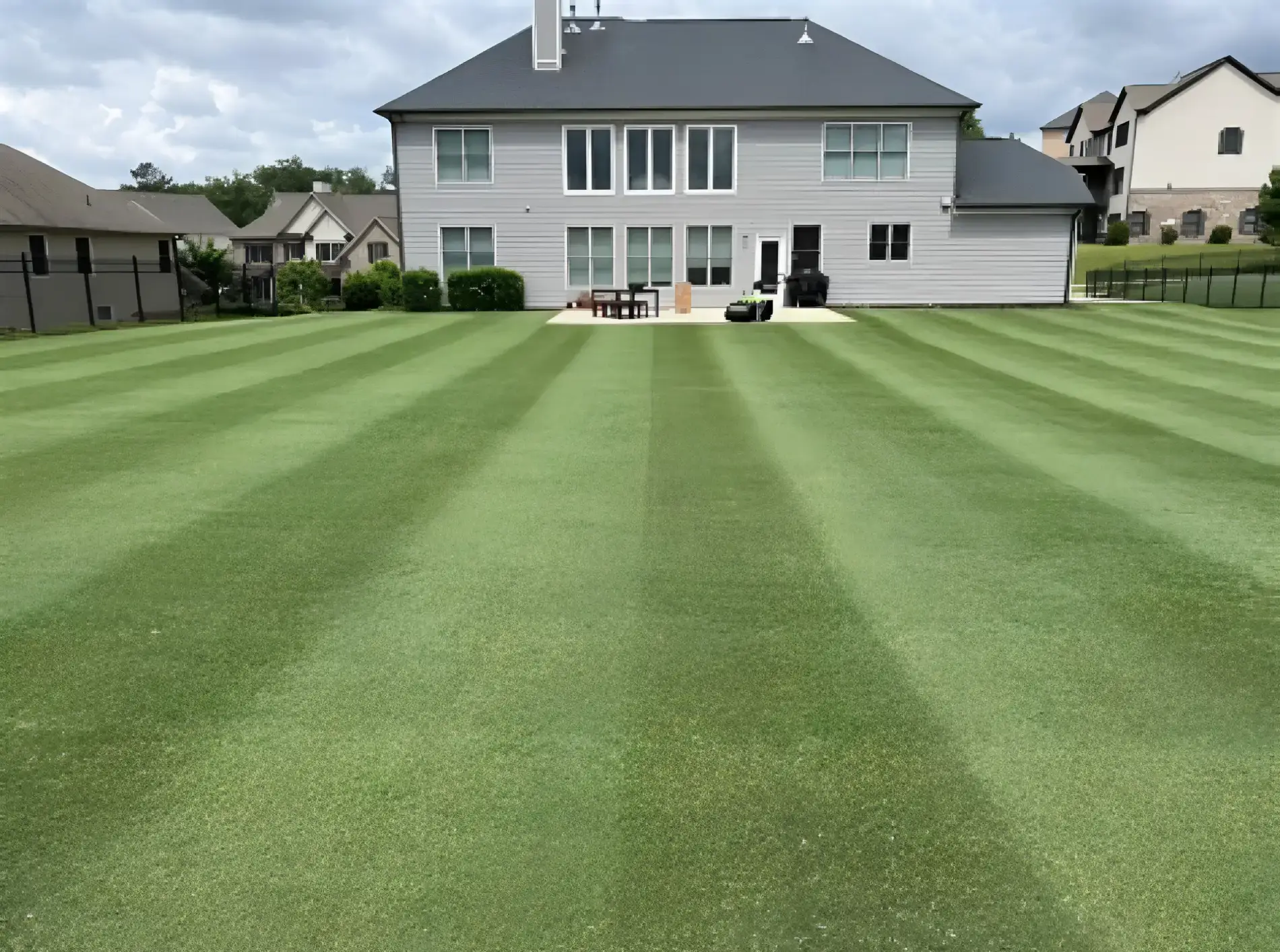 Large residential backyard featuring crisp, professionally mowed lawn stripes leading to a grey two-story house by Clean Cutz.